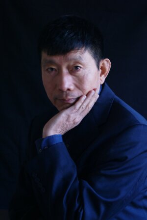 A photo of Dr. Tim Liao. He is standing in a dark blue shirt against a black background with his left hand resting on his cheek.