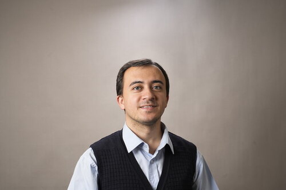 Yusuf Karipek stands against a neutral background. He wears a collared shirt and vest and is smiling slightly.