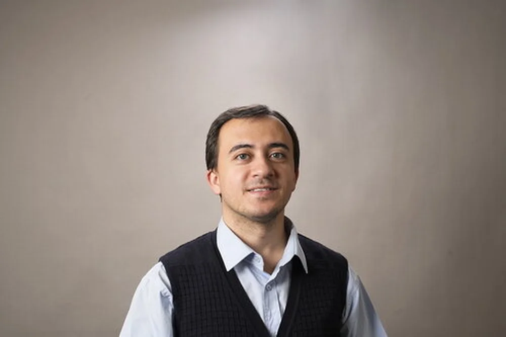 Yusuf Karipek stands against a neutral background. He wears a collared shirt and vest and is smiling slightly.