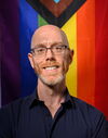 A photo of Dr. Stefan Vogler. He is standing in front of a Progress Pride flag and smiling at the camera.