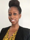 Nikel A. Bailey smiles at the camera. She wears a black blazer and yellow shirt and has her hair in a loc bun atop her head.