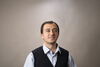 Yusuf Karipek stands against a neutral background. He wears a collared shirt and vest and is smiling slightly.