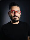 Ghassan Moussawi stands against a dark background. He wears all black clothing, except for red glasses.
