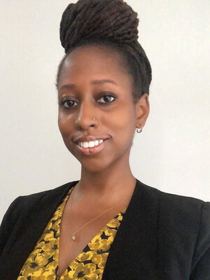 Nikel A. Bailey smiles at the camera. She wears a black blazer and yellow shirt and has her hair in a loc bun atop her head.