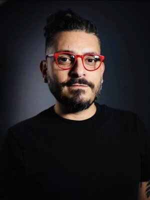 Ghassan Moussawi stands against a dark background. He wears all black clothing, except for red glasses.
