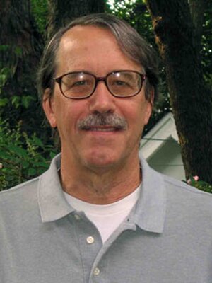 This is a photo of Douglas Barnes. He is wearing a grey polo shirt and standing in front of green foliage.
