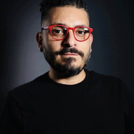 Ghassan Moussawi stands against a dark background. He wears all black clothing, except for red glasses.