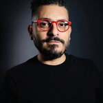 Ghassan Moussawi stands against a dark background. He wears all black clothing, except for red glasses.